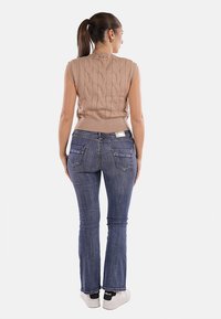 Maglione senza maniche in beige con motivo a treccia, abbinato a jeans flare in denim blu scuro, con tasche posteriori e un design aderente.