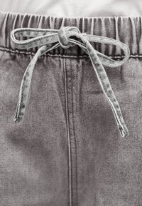 Εγγύς πλάνο γκρι τζιν παντελονιού με ελαστική μέση και δεμένο φιόγκο από ύφασμα στη μέση μπροστά.