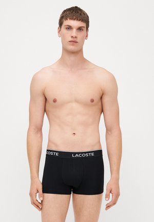 Shorts en coton noir avec une taille blanche arborant le logo "LACOSTE". Le tissu semble lisse et ajusté au corps du modèle.