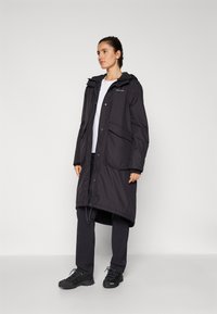 Schwarze lange Regenjacke mit Kapuze, Druckknopfverschluss, zwei Fronttaschen und einem leicht verlängerten hinteren Saum, getragen über dunklen Hosen und Schuhen.