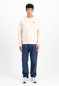 Creme sweatshirt med broderet logo, ribbet manchetter og kant; parret med blå denim jeans og sølvfarvede sportsko.