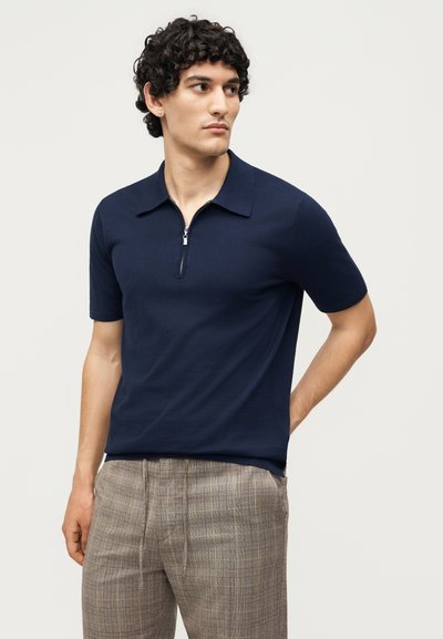 Άνδρας με σγουρά μαλλιά που φοράει σκούρο μπλε πόλο με κοντά μανίκια και φερμουάρ στο μπροστινό μέρος και καρό καφέ παντελόνια, κοιτάζοντας αριστερά σε απλό φόντο.