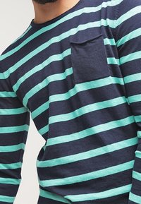 Pitkähihainen paita, jossa on vaakasuorat tummansinisen ja sinivihreän raidat, pyöreä kaula-aukko ja rintatasku, pehmeällä puuvillaisella koostumuksella.