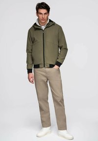 Mężczyzna w oliwkowej kurtce z kapturem, beżowych spodniach i białych sneakersach, stoi z jedną ręką w kieszeni na jednolitym tle.