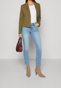 Žena v olivovém blejzru, bílém topu, světlých modrých džínách, béžových vysokých botách a držící červenou kostkovanou kabelku u boku.