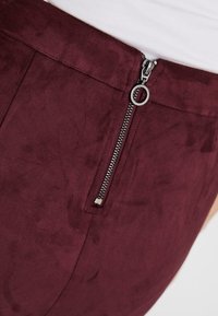 Burgunderfarbenes Faux-Suede-Gewebe mit einem versteckten Reißverschlussverschluss, der einen Metallzug mit glatter Oberfläche hat. Das Design ist tailliert und passgenau.