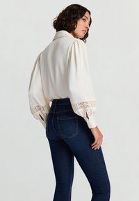 Cremefarbene Bluse mit Puffärmeln, Spitzenbesatz an den Manschetten, kombiniert mit dunkelblauen, hoch taillierten Jeans. Glatte Textur, klassisches Kragendesign.