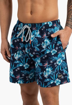Homme portant un short de bain bleu marine avec un imprimé floral tropical bleu et blanc, taille élastique et cordon blanc, main dans la poche.