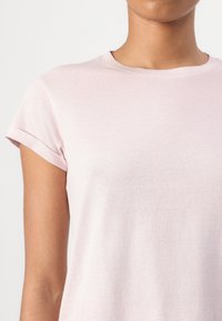 Prim-plan cu o persoană care poartă un tricou roz deschis, cu mânecă scurtă și decolteu rotund, cu tiv întors la mâneca stângă.
