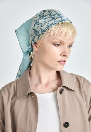 Foulard en soie avec un motif bleu et crème, comportant des motifs circulaires. Noué à l'arrière, avec une texture lisse. Le modèle porte une veste beige et un haut blanc.