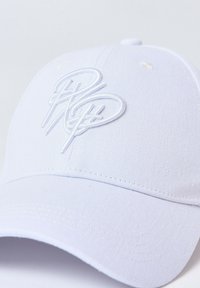 Gorra blanca con logo bordado, hecha de un tejido con textura suave. Presenta visera curvada y agujeros de ventilación en la parte superior.