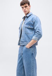 Νεαρός άνδρας που φοράει γυρισμένο μπουφάν τζιν και τζιν παντελόνι, στέκεται με τα χέρια στις τσέπες μπροστά σε απλό φόντο.