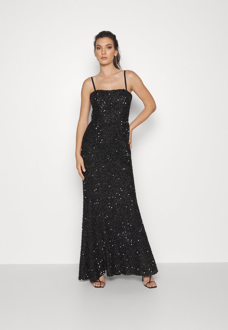 Donna in piedi contro uno sfondo bianco, indossa un lungo abito da sera nero con paillettes, spalline sottili e tacchi neri con cinturini.
