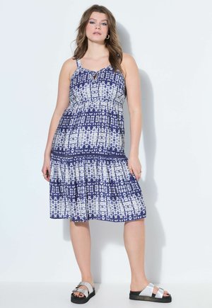 Kvinde iført ærmeløs blå og hvid mønstret knælængde kjole og sølvfarvede sandaler, stående mod en ensartet lys baggrund.