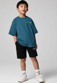 Tyrkis oversized t-shirt med gul logo, sorte shorts, hvide sneakers og hvide sokker. Glat tekstur og afslappet design.