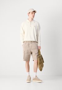 Polo bege de manga longa com três botões, calções cargo cor camelo, ténis claros e um boné bege; carregando uma mala verde.