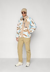 Giacca in fleece blu, marrone e bianco con motivi astratti, dotata di taschino beige e zip, abbinata a pantaloni e scarpe beige chiaro.