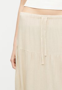 Jupe beige taille haute avec détail de ceinture à cordon, portée par une personne avec un haut blanc court laissant apparaître le ventre, la main gauche détendue.