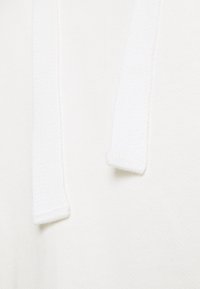 Bílá mikina z jemné tkaniny, která má měkkou texturu a ozdobné šňůrky s žebrovanými konci, což zdůrazňuje minimalistický a čistý design.