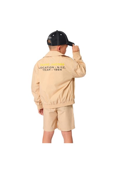 Enfant portant une veste et un short beige, tenant une casquette noire, dos tourné, avec le texte « MARC JACOBS LOCATION : N.Y.C. YEAR : 1984 » sur la veste.