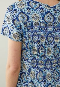 Modrá blůza se vzorem obsahujícím květinové a geometrické prvky, s nařaseným šitím na zadní straně a krátkými rukávy s háčkovaným lemem.