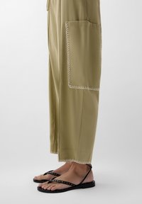 Khaki weite Hosen mit weißen dekorativen Nähten, ausgestattet mit zwei Seitentaschen. Schwarze flache Sandalen mit metallischen Nieten und feinen Riemen.