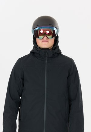 Personne portant une veste de ski noire, un casque noir et des lunettes de ski réfléchissantes, faisant face vers l'avant sur un fond blanc.