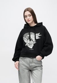 Mustast pehmest kangast kapuutsiga dressipluus, millel on valge graafilise koljumotiiv. Varustatud nööriga kapuutsiga ja soonikkoes varrukaservadega. Sobitatud heledate hallide teksadega.