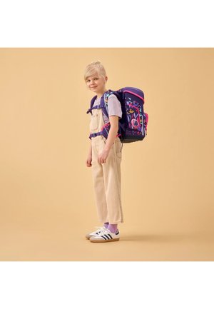 Junges Mädchen mit blondem Haar, das beige Overalls, ein lila Shirt und einen bunten Rucksack trägt, steht vor einem schlichten beigen Hintergrund.