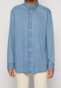 Camicia in denim blu chiaro con bottoni e maniche lunghe indossata sopra pantaloni beige e bianchi a fantasia visibili dalla vita in giù.