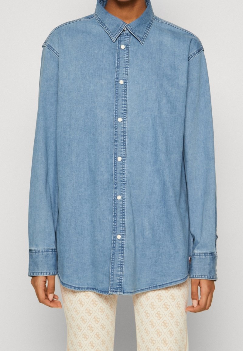 Camicia in denim blu chiaro con bottoni e maniche lunghe indossata sopra pantaloni beige e bianchi a fantasia visibili dalla vita in giù.
