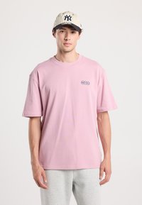 Pink short-sleeve t-shirt with round neckline and an embroidered "NYC" logo. Worn with gray sweatpants and a beige baseball cap featuring pinstripes.