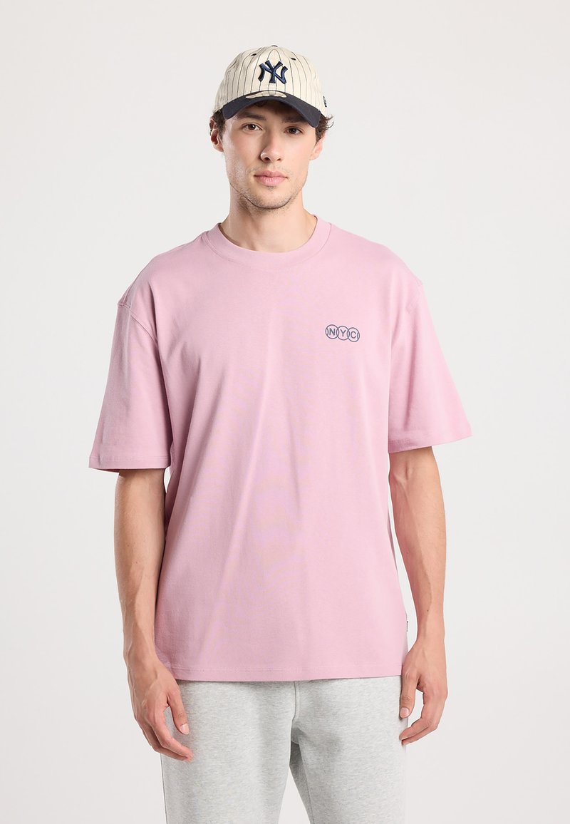 Pink short-sleeve t-shirt with round neckline and an embroidered "NYC" logo. Worn with gray sweatpants and a beige baseball cap featuring pinstripes.