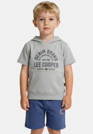 Garçon blond portant un sweat à capuche gris à manches courtes avec le texte "Lee Cooper" et un short bleu, debout devant un fond blanc.