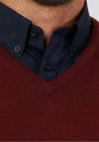 Burgunderfarbener V-Ausschnitt-Pullover über einem dunkelblauen Hemd mit Kragen und feinem Muster; sichtbare Knöpfe und Texturdetail im Stoff.