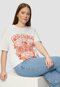 Witte oversized t-shirt met oranje bloemenafbeelding en tekstontwerp. Lichtblauwe jeans zichtbaar, met een gladde textuur en een ontspannen pasvorm.