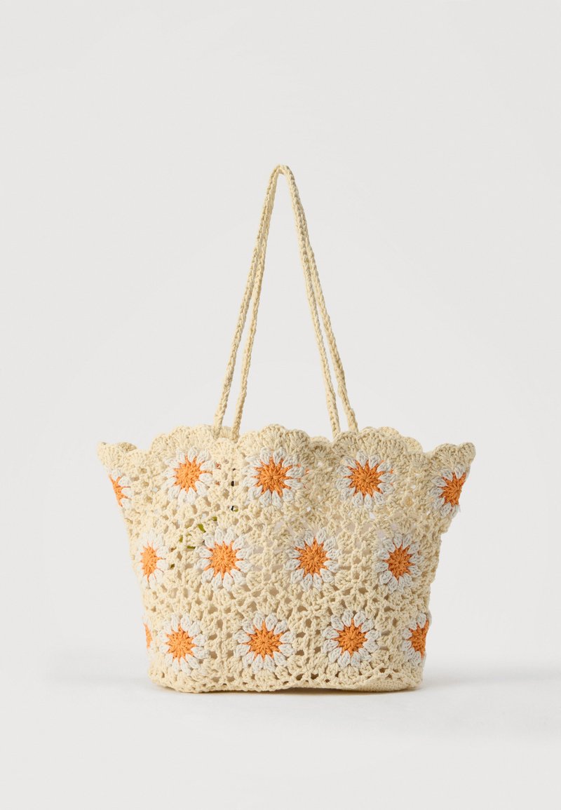 Sac fourre-tout beige crocheté avec motifs de marguerites orange et blanches, bords festonnés et deux longues poignées tressées.