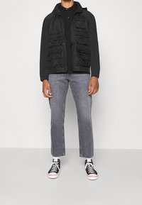 Homme portant une veste noire à poches multiples, une chemise noire, un jean gris et des baskets montantes noires avec lacets blancs, se tenant devant un fond blanc.