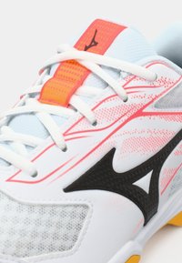 Białe buty sportowe z siatkową teksturą, czarnymi i różowymi akcentami oraz błyszczącym wykończeniem. Posiadają białe sznurówki i jaskrawopomarańczową etykietę.