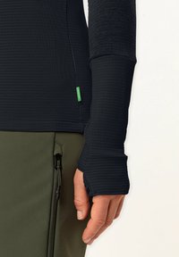 Schwarzes Langarmoberteil mit strukturiertem Muster und Daumenlöchern. Kombiniert mit olivgrünen Hosen, die eine sichtbare Reißverschlusstasche haben.