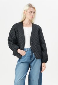 Casaco preto acolchoado com um ajuste descontraído, apresentando um decote em V, dois botões e um bolso frontal. Usado sobre uma blusa branca e jeans azuis.