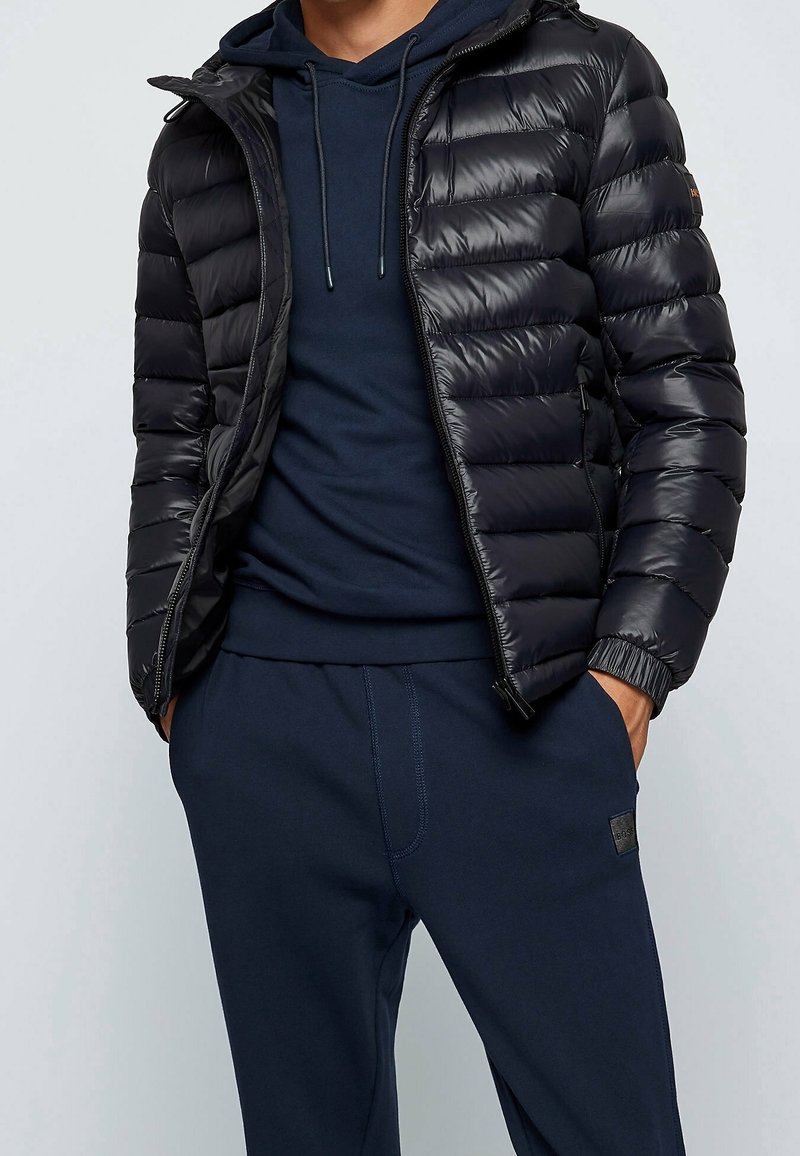 Homme portant une doudoune noire par-dessus un sweat à capuche bleu foncé avec un pantalon de jogging bleu foncé assorti, mains dans les poches, debout devant un fond clair.