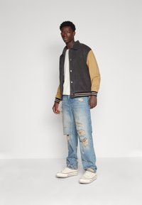 Veste varsity noire et beige avec des boutons pression, poignets côtelés et jeans droits bleu clair déchirés. Baskets beiges avec semelle blanche épaisse.