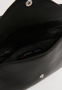 Interno di una borsa a tracolla in pelle nera con una texture liscia, dotata di una chiusura a scatto in argento e un'etichetta interna con "even & odd".