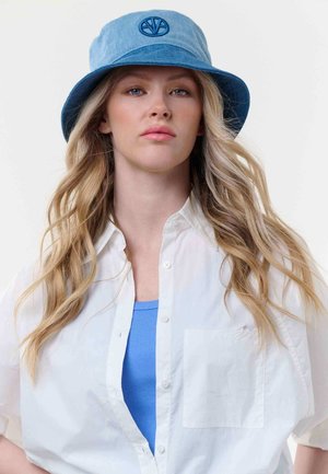 Junge Frau mit langem blondem Haar trägt einen blauen Anglerhut, ein weißes Hemd über einem blauen Top und blickt mit neutralem Gesichtsausdruck geradeaus.