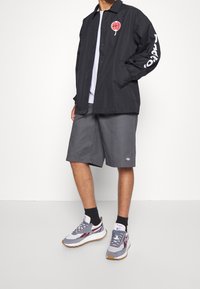 Svart knäppjacka med röd grafik, grå shorts och grå sneakers med detaljer. Enkel design med avslappnad passform.