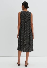Schwarzes ärmelloses Kleid mit fließender Silhouette, verfügt über ein weißes Punktenmuster, runden Ausschnitt und eine Kellenlänge. Dezente Textur im gesamten Kleid.