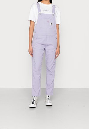 Vaaleanlilat overallsit, joissa on säädettävät hihnat, etutaskut ja metallisten napin korostuksia, yhdistettynä valkoiseen minimalistiseen t-paitaan ja kuviollisiin lenkkareihin.