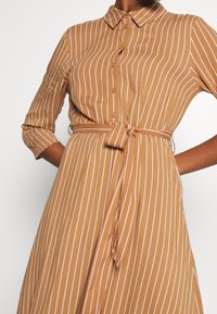 Vestido de camisa de rayas marrones con cuello, cierre de botones y cintura ajustada. Presenta rayas verticales blancas y mangas de tres cuartos.