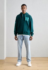Uomo in piedi al chiuso che indossa un felpa grafica teal, jeans azzurri chiari e sneaker bianche su un pavimento in legno con sfondo di parete grigia chiara e semplice.